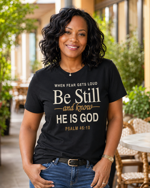 A smiling woman with dark curly hair stands outdoors wearing a black graphic T-shirt with the text 'WHEN FEAR GETS LOUD Be Still and know HE IS GOD PSALM 46:10' in white and gold lettering, paired with blue jeans.