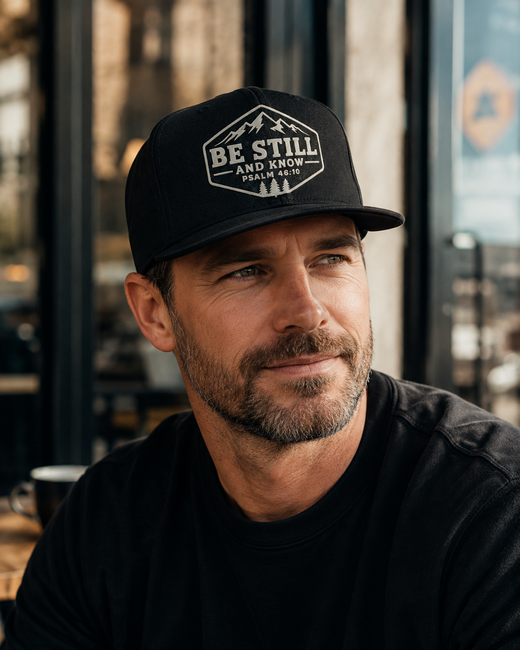 A man wearing a black baseball cap with a white graphic logo that reads 'BE STILL AND KNOW' with mountain and tree illustrations, photographed in a softly blurred indoor setting.