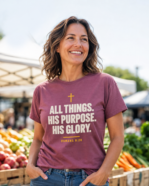 A smiling woman wearing a maroon graphic t-shirt with a yellow cross and the text 'ALL THINGS. HIS PURPOSE.