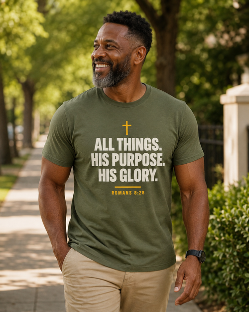 Smiling man wearing an olive green Christian T-shirt reading “All Things. His Purpose. His Glory.” with Romans 8:28 while walking outdoors.