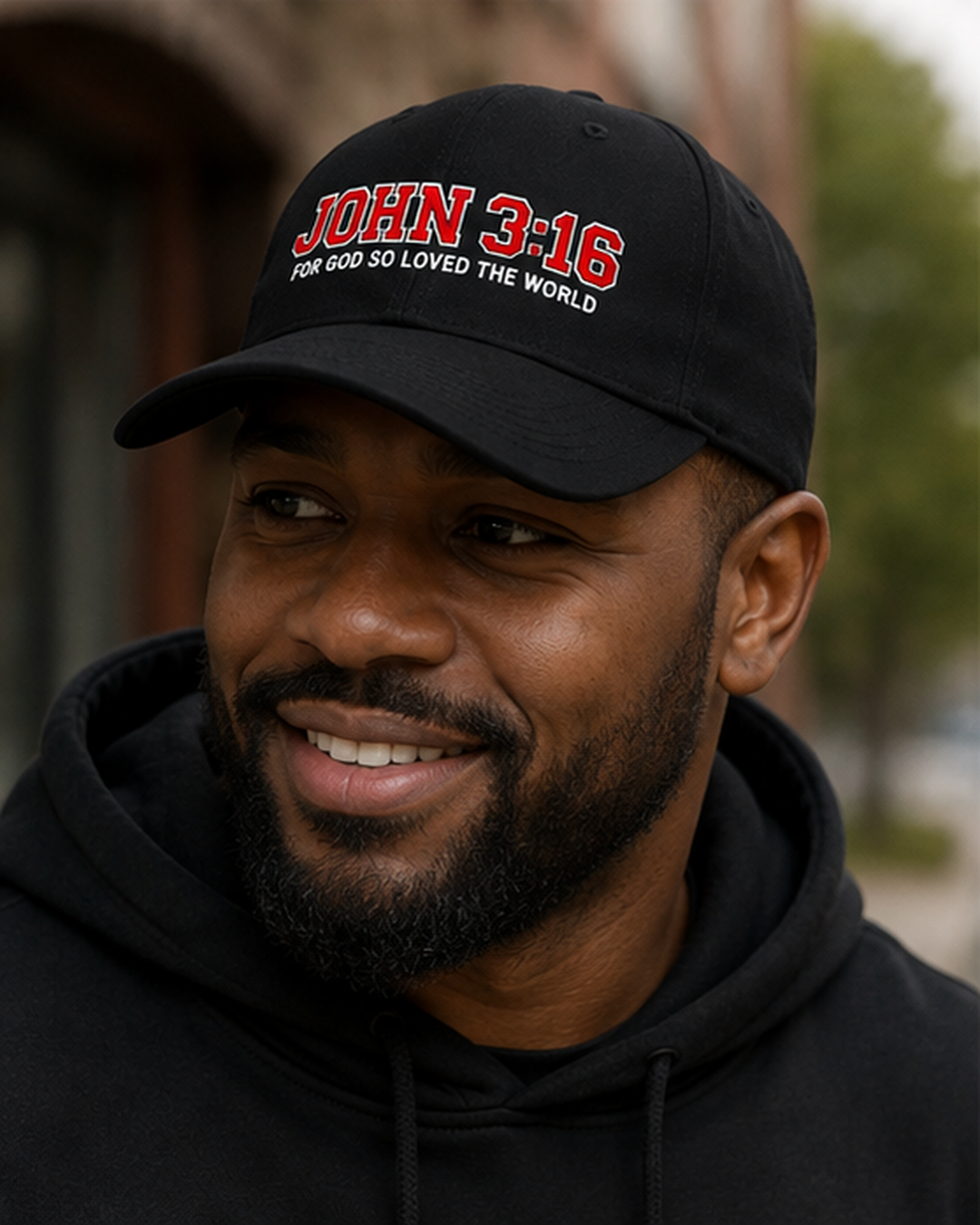 A smiling man wearing a black baseball cap with red and white embroidered text reading 'JOHN 3:16 FOR GOD SO LOVED THE WORLD', photographed outdoors with a blurred background.