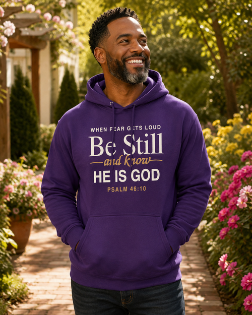 A smiling man wearing a purple hoodie with a Bible verse graphic on the chest, standing in a garden pathway surrounded by flowers and greenery.