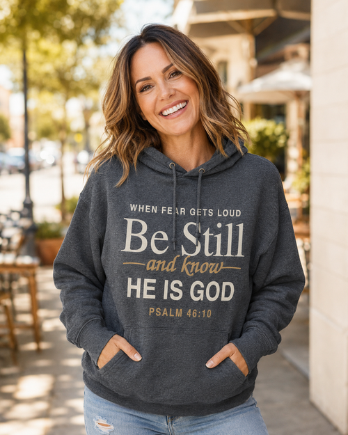 A woman smiling outdoors wearing a dark heather gray hoodie with a white and gold Bible verse print on the front, paired with light blue jeans.