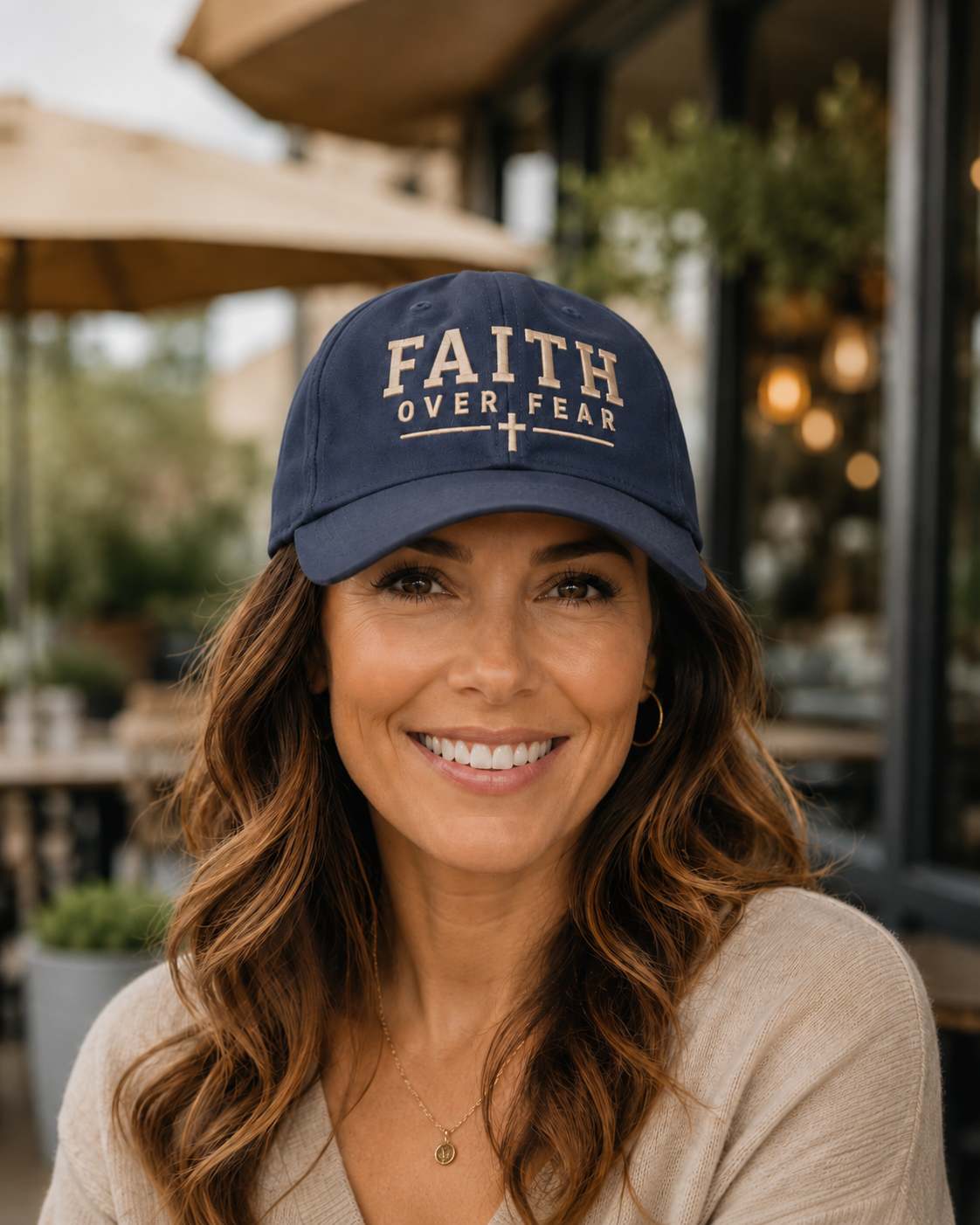 A smiling woman with wavy brown hair wearing a navy blue baseball cap with 'FAITH OVER FEAR' embroidered in beige on the front, photographed in an outdoor cafe setting.